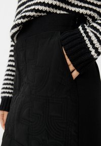 Schwarzer gesteppter Rock mit tonal abgestimmten Stickmustern und seitlichen Taschen. Kombiniert mit einem gestreiften, grob gestrickten Pullover in Schwarz und Weiß.