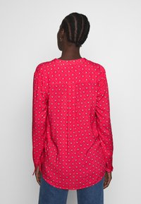Camicia rossa a maniche lunghe con un motivo a rombi blu e bianchi, dotata di un orlo arrotondato e dettagli sulla cucitura posteriore. Realizzata in tessuto leggero.