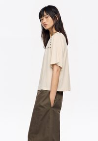 Top beige de manga corta con un corte relajado, que presenta adornos de perlas en el escote. Combinado con pantalones verde oscuro, con bolsillos laterales visibles.