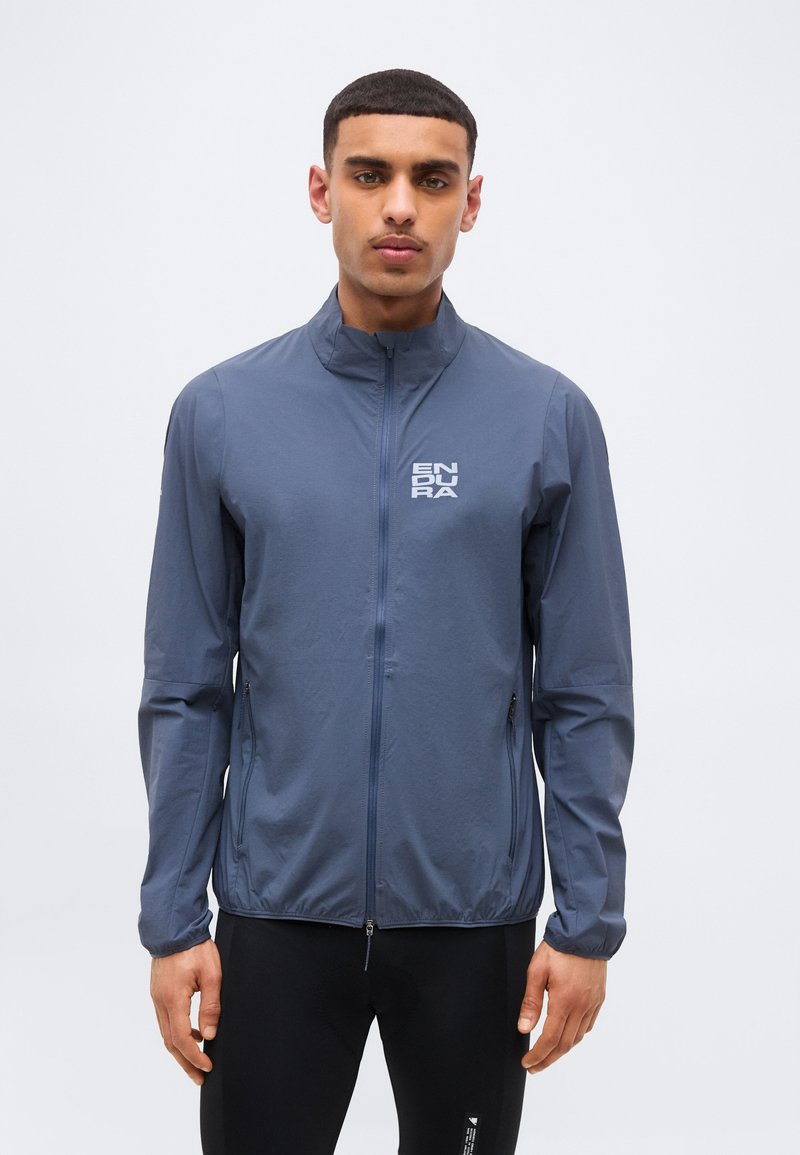 Άνδρας που φοράει μπλε ελαφρύ μπουφάν με φερμουάρ και το λογότυπο "ENDURA" στο στήθος, μαζί με μαύρο παντελόνι, μπροστά σε απλό φόντο.