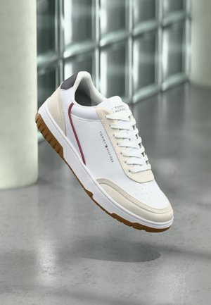 Biały i beżowy sneaker Tommy Hilfiger z gumową podeszwą, czerwonym akcentem i firmowym napisem, unoszący się nad polerowaną betonową podłogą.
