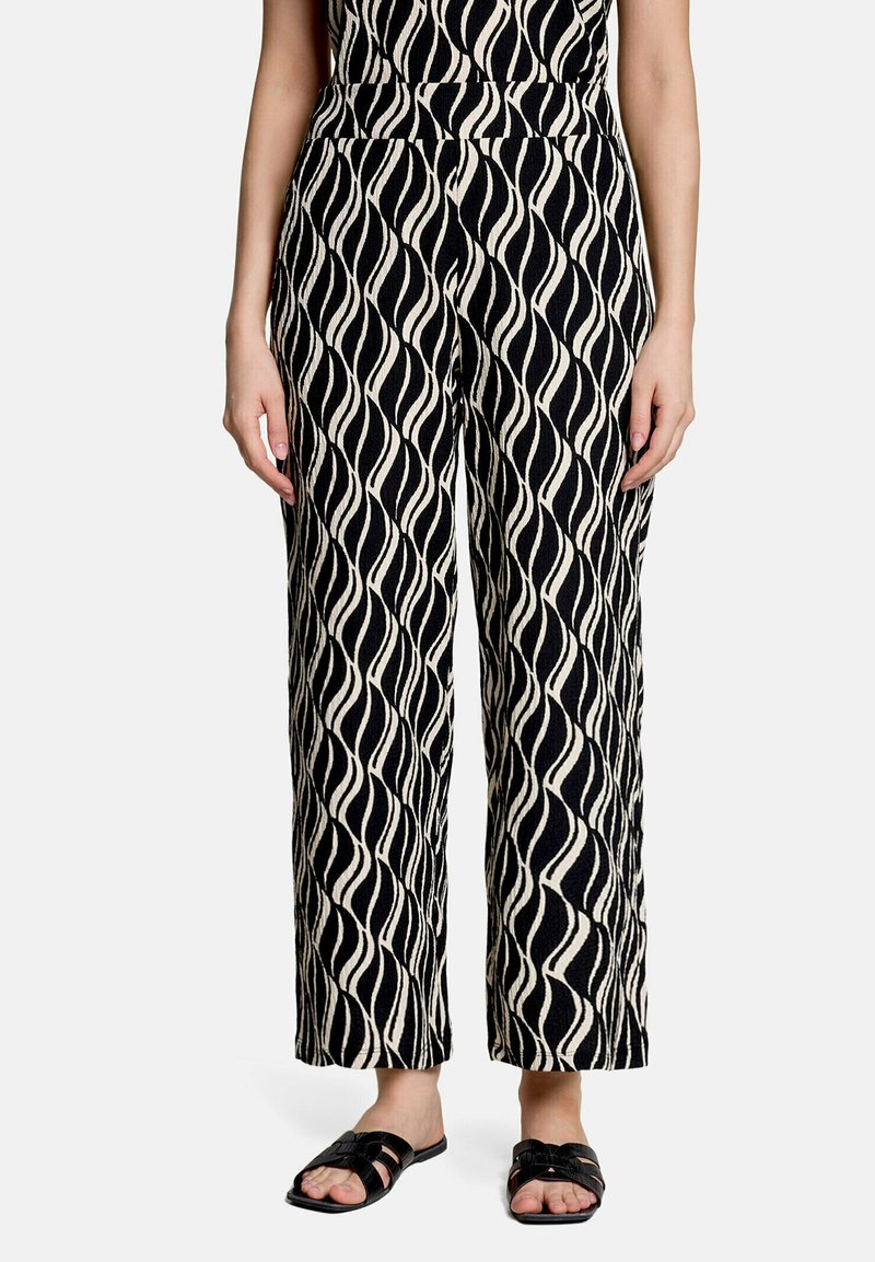 Femme portant un pantalon large à motif ondulé noir et blanc et des sandales plates noires avec les orteils découverts, debout devant un fond uni.