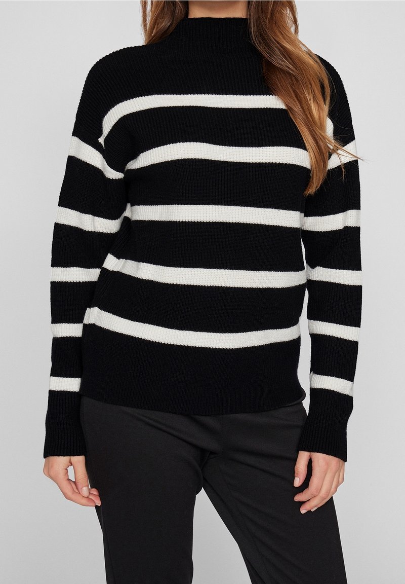 Femme portant un pull en maille noir avec des rayures blanches horizontales et un pantalon noir, debout devant un fond gris clair uni.