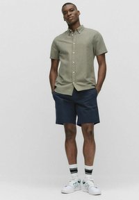 Olijfgroen overhemd met korte mouwen en knopen, gecombineerd met marineblauwe shorts. Witte sneakers met groene accenten en gestreepte sokken maken de outfit compleet.