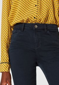 Pantalones azul marino con una textura suave, que cuentan con bolsillos delanteros, un cierre de botón metálico y detalles de costura visibles. Combinados con una camisa amarilla con estampado.
