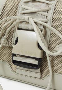 Šviesiai rudos spalvos sportbačiai su tinklelio detalėmis, plokščiomis raišteliais ir išskirtine plastiko sagtimi su "STEVE MADDEN" logotipu. Lygios odos akcentai.