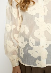 Lekka, prześwitująca kremowa bluzka z dużym kwiatowym wzorem; posiada guziki z przodu, bufiaste rękawy i teksturowane akcenty.