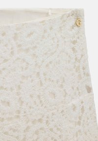 Tissu en dentelle blanche avec des motifs floraux au crochet et un petit pendentif doré en forme de lettre G cousu près du bord supérieur.