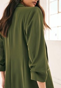 Blazer verde oliva con un corte relajado, mangas largas y puños enrollados. Tejido suave con un sutil brillo, que presenta un detalle en la costura trasera.