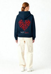 Sweat à capuche marine avec un design de cœur rouge composé de plusieurs petits cœurs, avec le texte "SWEET heart" en dessous. Assorti avec un pantalon cargo crème.