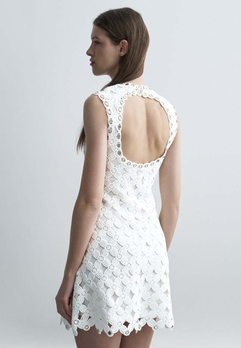 Γυναίκα με λευκό αμάνικο φόρεμα από δαντέλα, με μεγάλο ανοιχτό πίσω μέρος και κυκλικά κοψίματα, που στέκεται μπροστά σε απλό ανοιχτό γκρι φόντο.