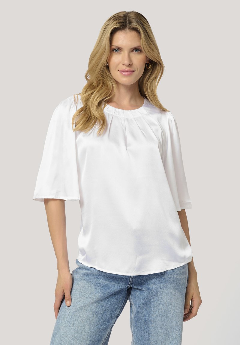 Haut en satin blanc avec des manches courtes et larges et un col arrondi, mettant en valeur de douces plis aux épaules et une coupe décontractée.