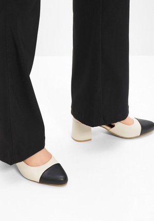 Scarpe con tacco beige e nere a due tonalità, con punta rotonda e tacco a blocco, abbinate a pantaloni neri, che mostrano una consistenza liscia.