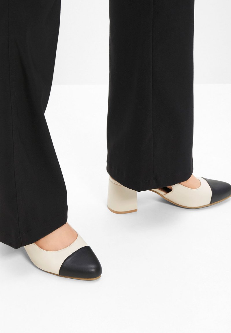 Scarpe con tacco beige e nere a due tonalità, con punta rotonda e tacco a blocco, abbinate a pantaloni neri, che mostrano una consistenza liscia.