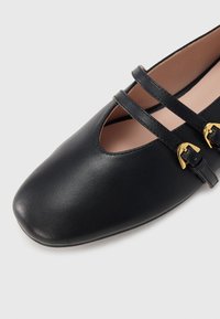 Μαύρα δερμάτινα παπούτσια με στρογγυλεμένη μύτη, διακοσμημένα με δύο σταυρωτές λουρίδες και χρυσά αγκράφες. Λείο υλικό με σχέδιο κοψίματος κοντά στη μύτη.