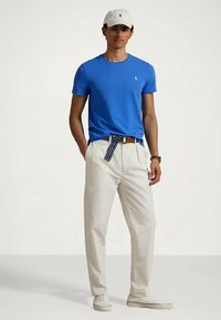 Άνδρας ντυμένος με μπλε κοντομάνικο πουκάμισο, λευκά παντελόνια με ριγωτή ζώνη, λευκά αθλητικά παπούτσια, λευκό καπέλο και ρολόι χειρός, στέκεται με τα χέρια στις τσέπες.