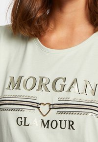 Camiseta de algodón verde claro con letras metálicas plateadas que dicen "MORGAN" y "GLAMOUR", con un acento en forma de corazón y un patrón de rayas.
