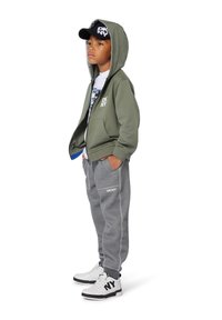 Sweat à capuche vert olive avec fermeture éclair, pantalon de survêtement gris, t-shirt graphique blanc, casquette noire avec "DKNY", et baskets noir et blanc.