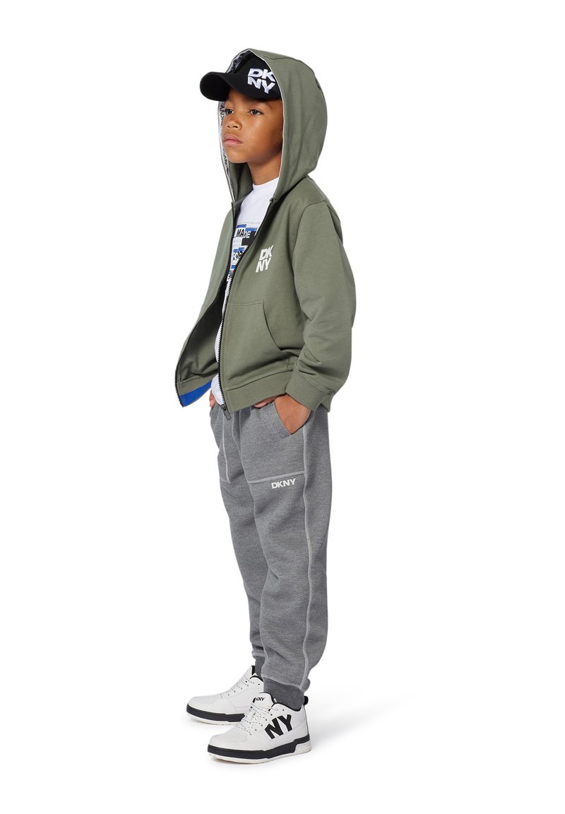 Sweat à capuche vert olive avec fermeture éclair, pantalon de survêtement gris, t-shirt graphique blanc, casquette noire avec "DKNY", et baskets noir et blanc.