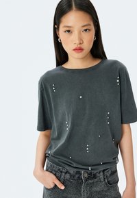 Camiseta gris de manga corta con piedras brillantes dispersas en el frente. Textura suave y corte relajado, combinada con jeans oscuros.