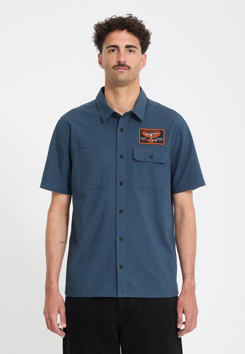 Homme portant une chemise bleue à manches courtes avec deux poches poitrine, dont une avec un patch Volcom Workwear noir et orange.