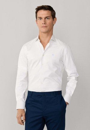 Homme portant une chemise blanche ajustée à boutons, rentrée dans un pantalon bleu foncé, debout avec une main dans la poche devant un fond uni.