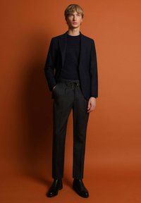 Blazer ajustado navy, suéter de punto negro y pantalones grises con cintura de cordón; el modelo lleva botas de cuero negras hasta el tobillo. Fondo naranja.