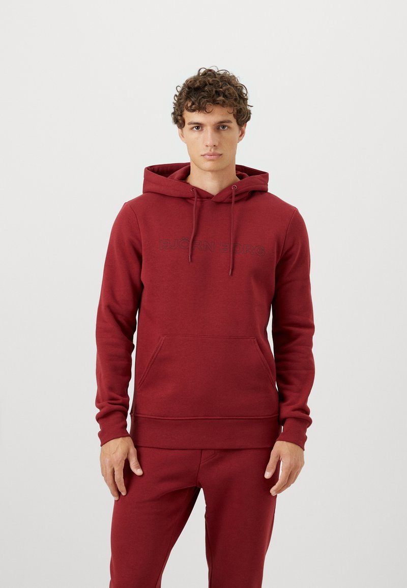 Mladý muž s kučeravými vlasmi v bordovom mikinovom kabáte a zodpovedajúcich teplákoch, stojí proti jednoduchému svetlému pozadí.