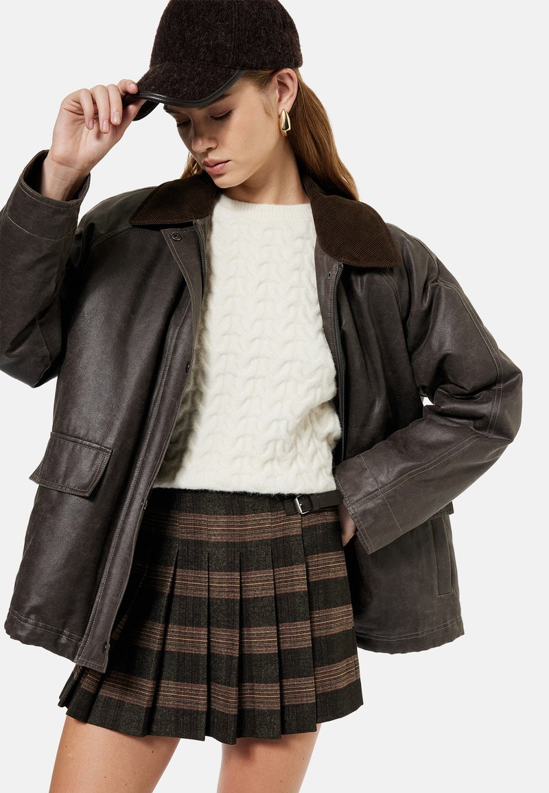 Giacca in pelle marrone con colletto in velluto a coste, maglione crema a trecce, e gonna a quadri con strisce marroni scure e chiare.