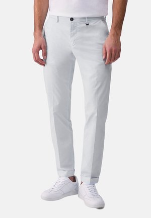 Uomo che indossa pantaloni slim fit grigio chiaro con risvolti arrotolati e sneakers bianche, in piedi davanti a uno sfondo chiaro e uniforme.