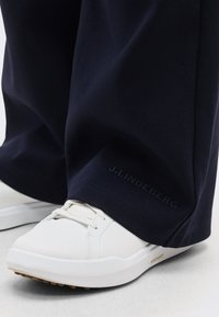 Λευκό αθλητικό παπούτσι με μπεζ σόλα με την ένδειξη "Archfit", φορεμένο με σκούρο μπλε παντελόνι που δείχνει την επωνυμία "J.Lindeberg" κοντά στον αστράγαλο.