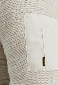 Gebreide crèmekleurige trui met een textuurpatroon, voorzien van een gladde beige stoffen zak en een zwart label dat aan de rand is gestikt.