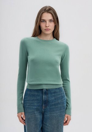Giovane donna con capelli lisci castano chiaro indossa un maglione verde chiaro a maniche lunghe e jeans blu a vita alta, in piedi davanti a uno sfondo semplice.