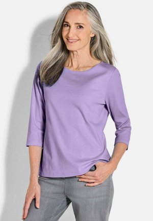 Glunderende vrouw van middelbare leeftijd met lang grijs haar, gekleed in een lavendelkleurig shirt met driekwart mouwen en grijze broek, staande met één hand in de zak.