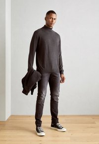 Mørk grå strik turtleneck, tætsiddende, parret med falmet sorte jeans og sorte canvas sneakers. En sort denimjakke holdes i venstre hånd.