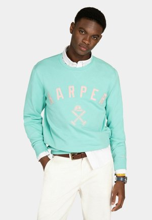 Hombre vistiendo una sudadera turquesa con el texto "HARPER" y mazos de polo cruzados, camisa blanca, pantalones blancos, cinturón marrón y reloj de pulsera.