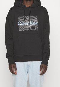 Felpa nera con cappuccio realizzata in morbido tessuto, dotata di una tasca frontale, caratterizzata da linee ondulate bianche e la scritta "Calvin Klein" in azzurro chiaro.