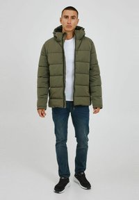 Padded green jacket with a hood, featuring a zip front and textured finish. Worn over a white shirt, paired with blue jeans and black sneakers.