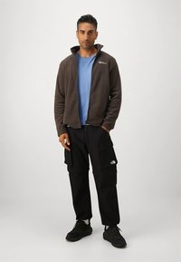 Hnědá fleecová bunda přes modrou košili, doplněná černými kalhotami typu cargo. Tenisky mají černý texturovaný materiál. Výrazné logo na bundě a kalhotách.