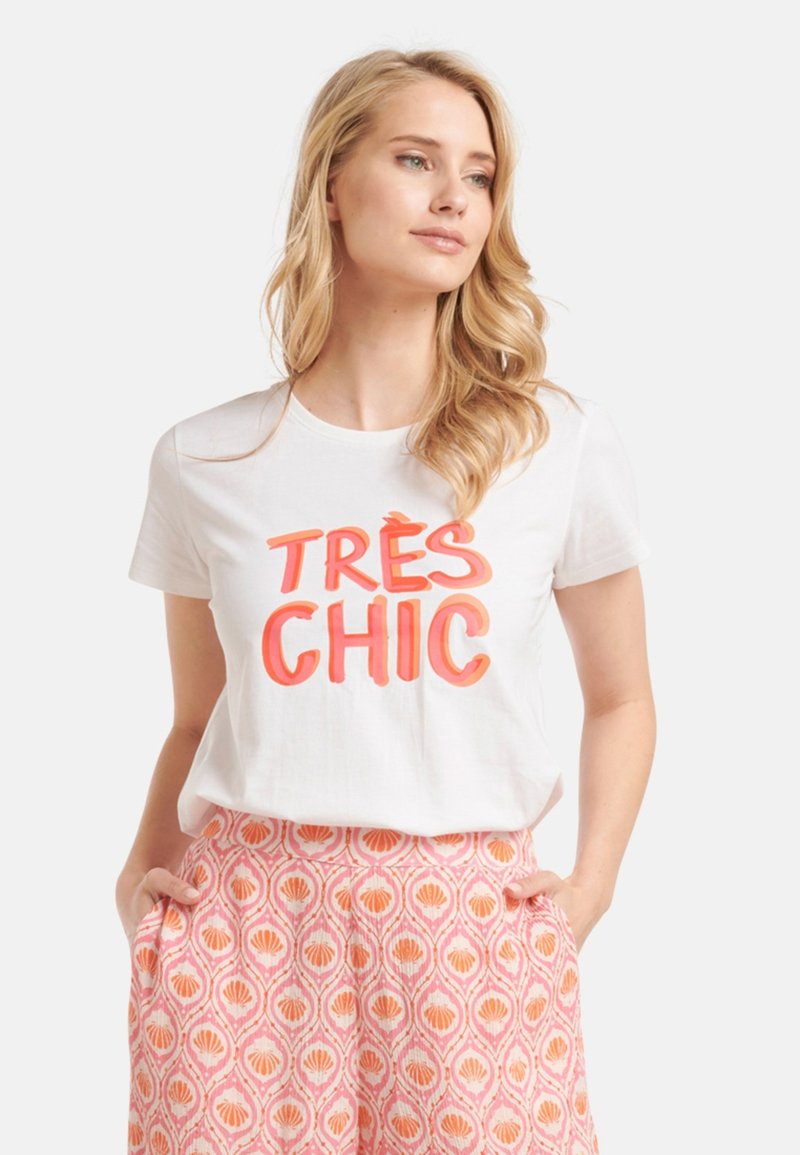 Weißes Kurzarm-T-Shirt mit fettem rot-orangefarbenem Text "TRÈS CHIC." Kombiniert mit gemusterten rosa Hosen mit Muschelmotiven.