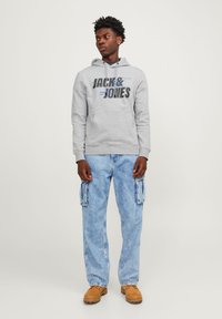 Sudadera gris con el logo de "JACK & JONES", combinada con pantalones cargo de mezclilla azul claro y botas color beige. Estilo informal con capucha ajustable y bolsillos laterales.