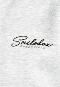 Un tessuto di cotone grigio chiaro presenta il ricamo di "Smilodox" in nero e "ESSENTIALS" in grigio, creando una consistenza morbida e un design minimalista.