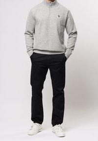 Grauer Reißverschluss-Pullover mit hohem Kragen und gesticktem Logo, kombiniert mit schwarzen Hosen und weißen Turnschuhen. Glatter Stoff mit entspannter Passform.