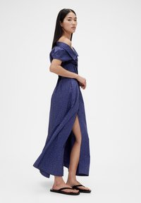 Abito lungo blu a pois bianchi con spalle scoperte, taglio aderente, spacco laterale, tessuto strutturato, indossato con infradito nere.