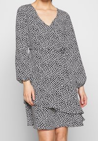 Black dress with a V-neck and long sleeves, featuring a white abstract pattern. The hem includes a ruffled detail and an adjustable waist tie.