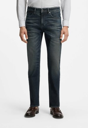 Mann trägt dunkelblaue Slim-Fit-Jeans, braune Lederschuhe und ein hell gestreiftes Hemd, das in die Hose gesteckt ist, vor weißem Hintergrund.