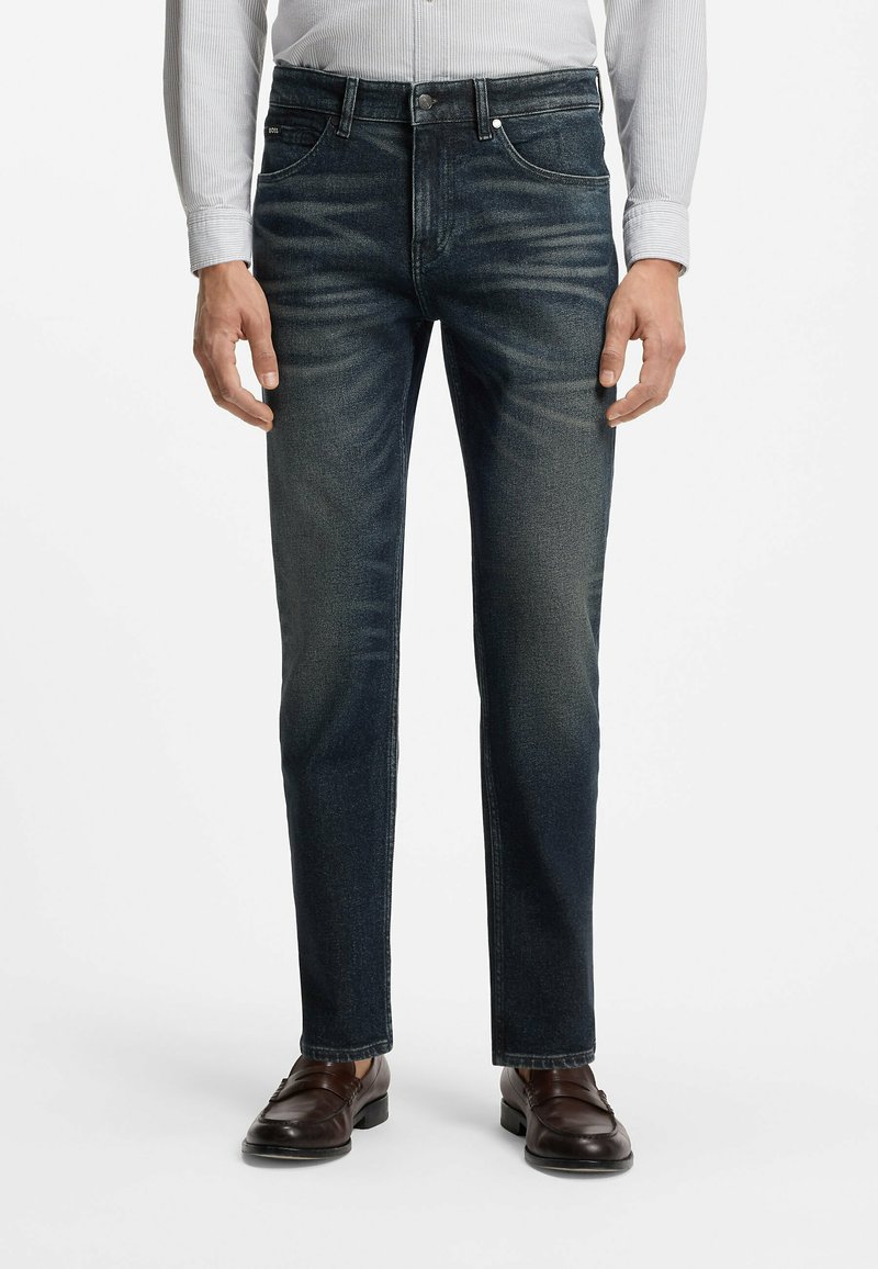 Homme portant un jean slim bleu foncé, des chaussures habillées en cuir marron et une chemise claire à rayures rentrée dans le pantalon sur fond blanc.