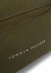 Borsa in nylon verde oliva con zip e finitura testurizzata. Presenta il marchio "TOMMY HILFIGER" in bianco con un accento a blocchi di colore.