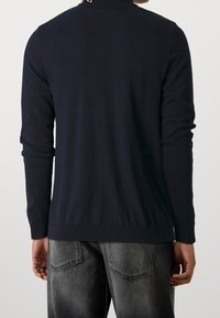 Man i en åtsittande svart långärmad polotröja och urtvättade svarta jeans, sedd bakifrån mot en enfärgad bakgrund.