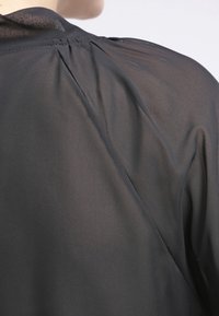 Černá, lehká blůza s průhlednou texturou, která má jemné plisování na ramenou a elegantní, štíhlý design.